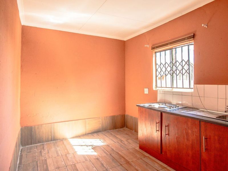A compact, empty kitchen featuring wooden cabinetry, a stainless steel sink, a built-in hob, and tiled flooring with a security-fitted window.