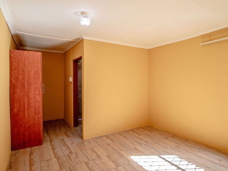 A simple, empty bedroom featuring tiled flooring, a built-in wardrobe, and a window fitted with security bars.