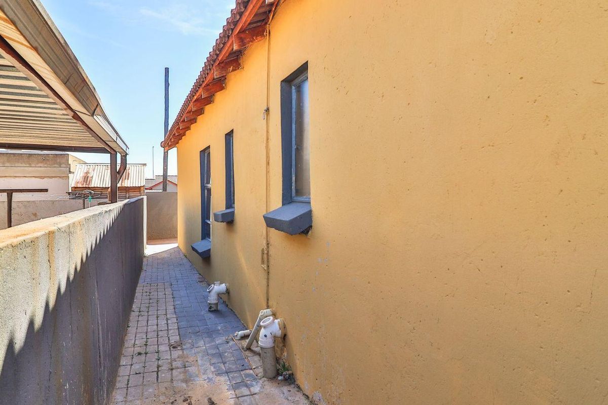 A side exterior view of the property featuring a paved walkway, boundary wall, and exposed exterior plumbing along the yellow rendered wall.