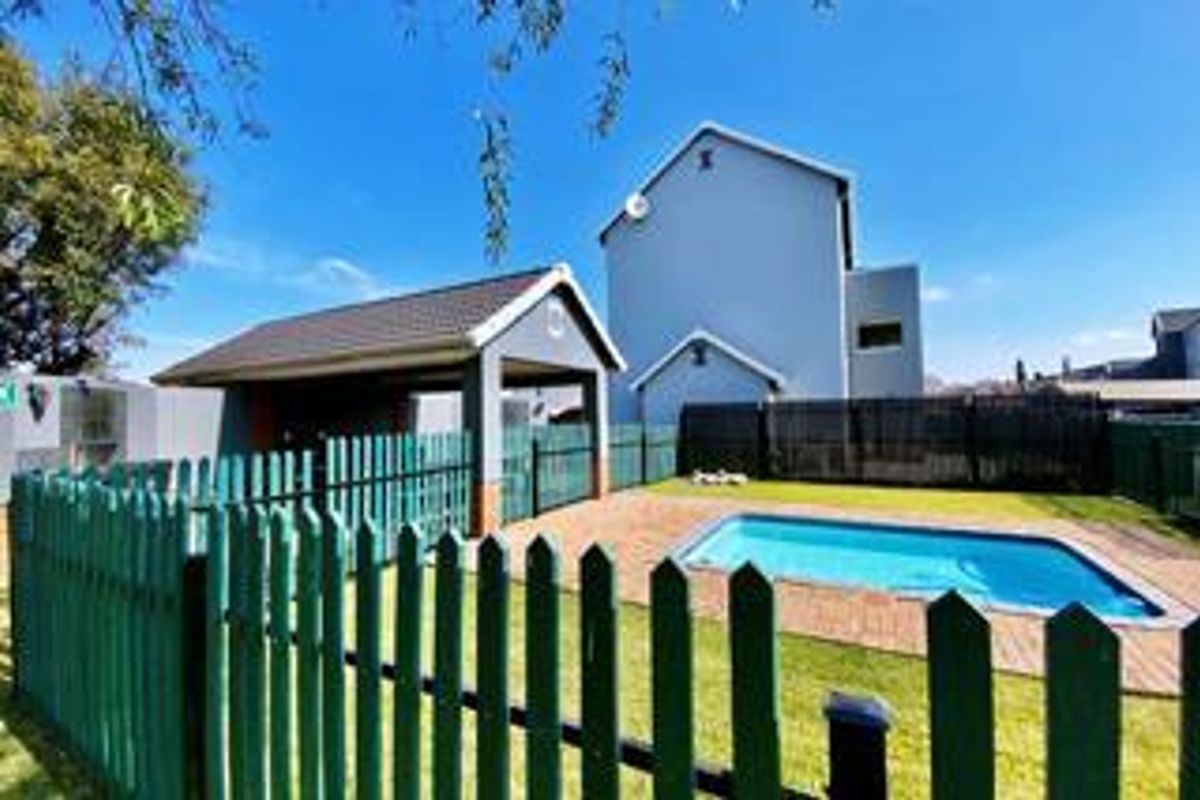 Rear exterior view featuring a private swimming pool, paved deck area, covered patio, and a well-maintained lawn enclosed by a picket fence.