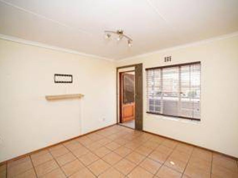 A bright, empty living room featuring neutral tiled flooring, a wall-mounted shelf, and large windows fitted with venetian blinds.
