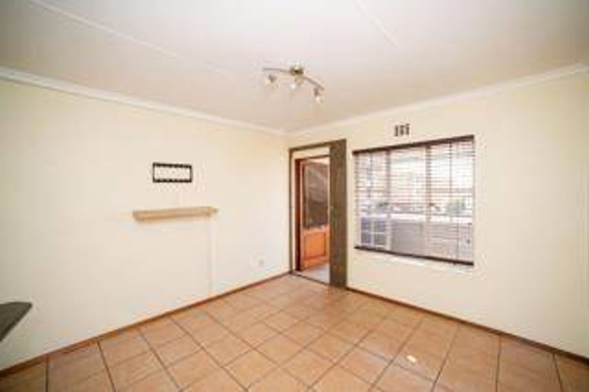 A bright, empty living room featuring neutral tiled flooring, a wall-mounted shelf, and large windows fitted with venetian blinds.