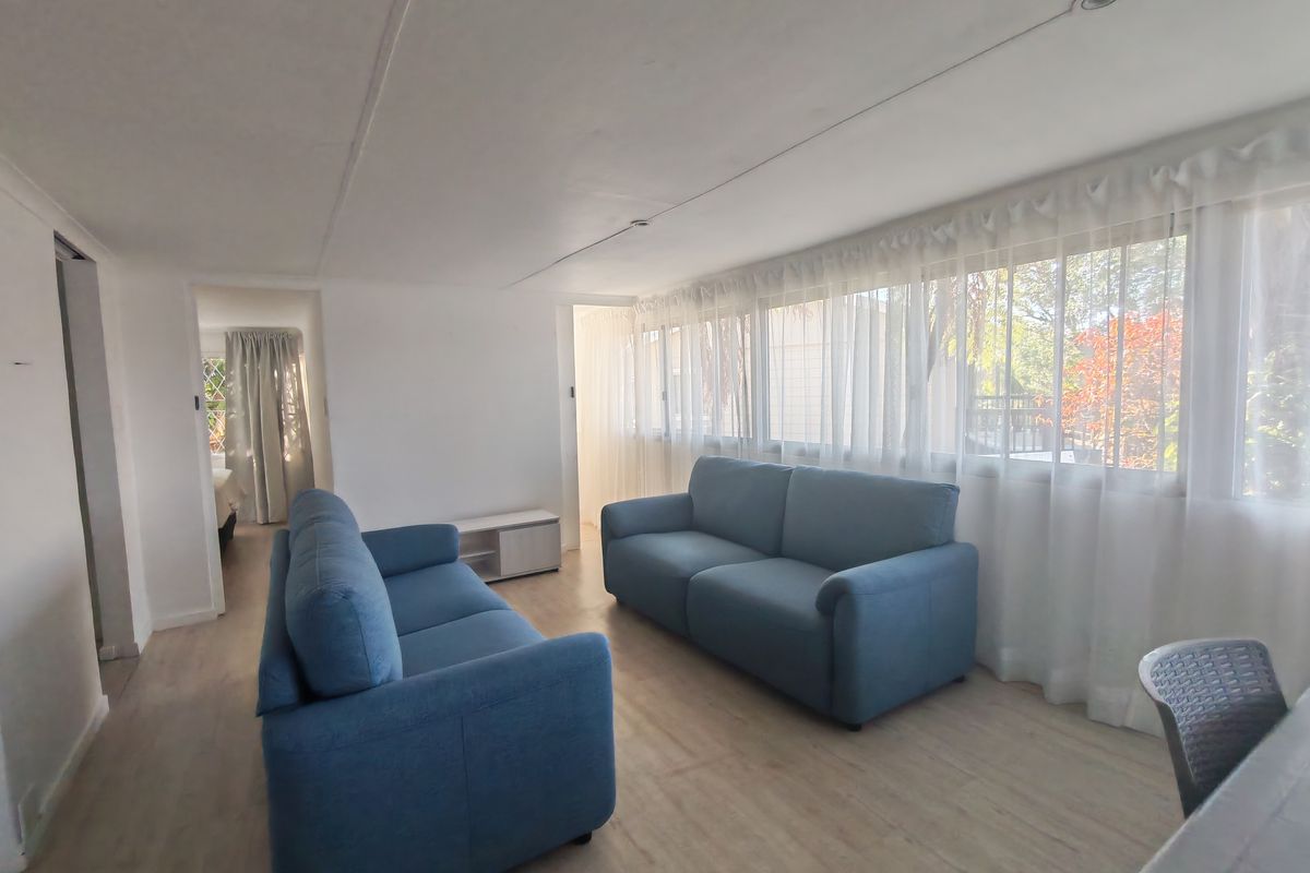 A bright living room featuring light-toned laminate flooring, large windows with sheer curtains, and recessed ceiling lighting, offering a clean and functional space.