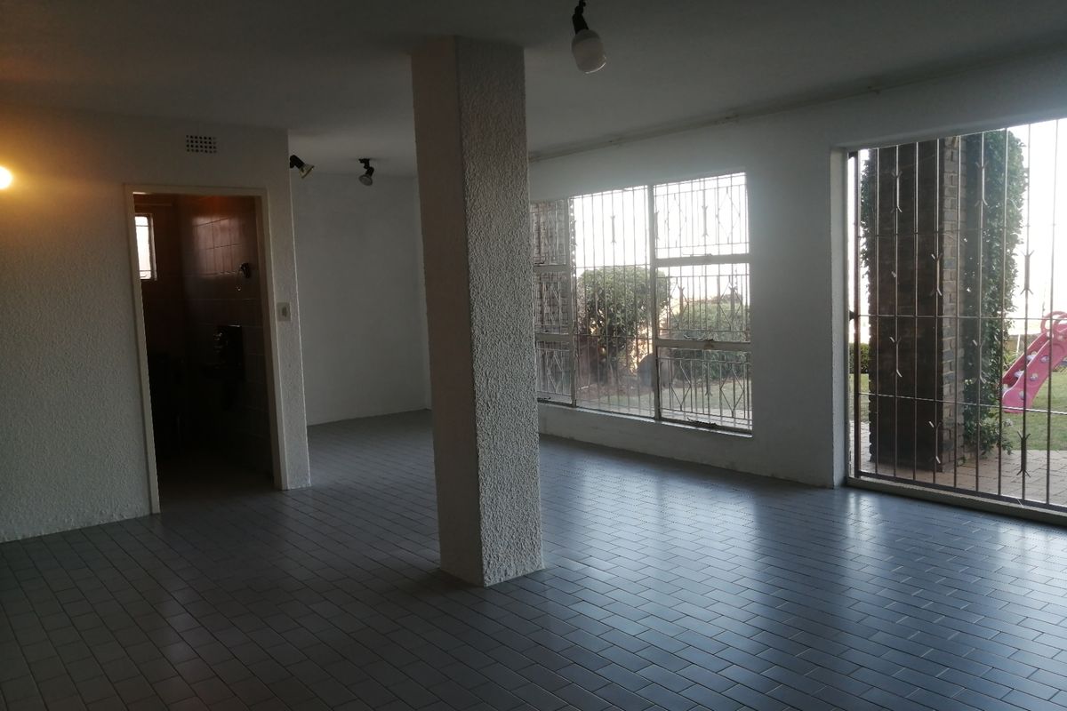 An empty, open-plan living area featuring tiled floors, a central structural column, and large windows with security bars, offering direct access to the garden.