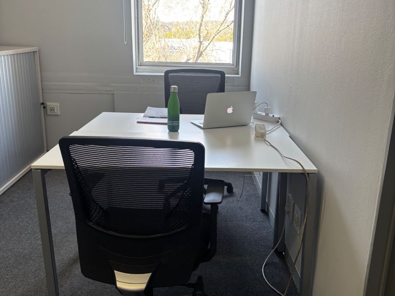 A functional office space featuring carpeted flooring, a window with a roller blind, and integrated electrical outlets.
