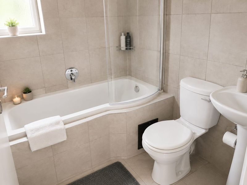 A clean, modern bathroom featuring a pedestal sink, toilet, and a bathtub with a glass shower screen and neutral wall tiling.