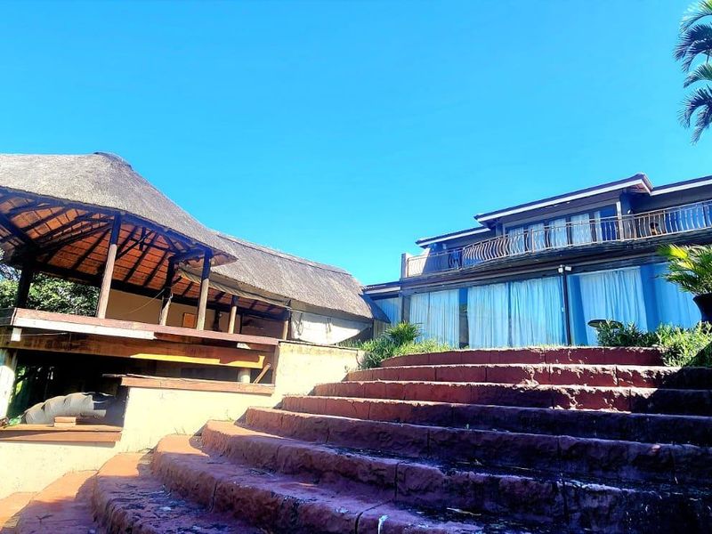 Rear exterior view featuring a thatched lapa, stone staircase leading to the main house, and a balcony with sliding glass doors.