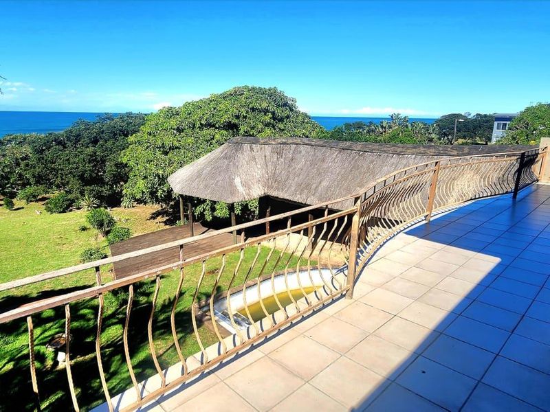 An elevated balcony overlooking a garden, swimming pool, and coastal view, featuring tiled flooring and a metal balustrade.