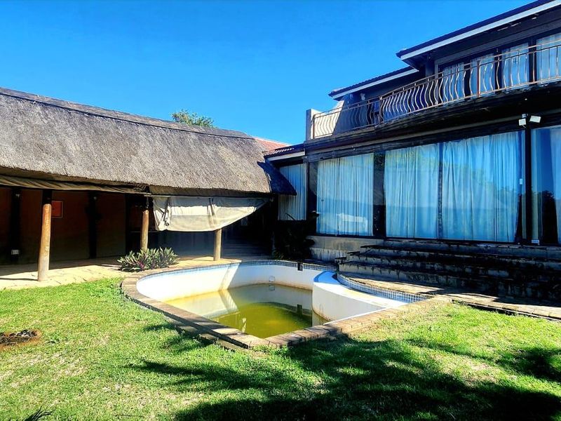 Outdoor entertainment area featuring a swimming pool, a thatched lapa, and a multi-story residence with a balcony and large glass sliding doors.