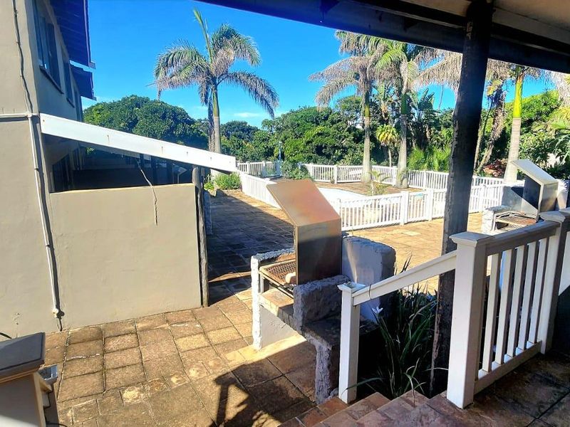 A covered outdoor patio area featuring a built-in braai, paved flooring, and white railings, overlooking a garden space.
