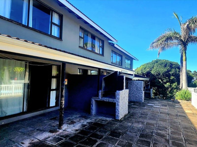 Rear exterior view featuring a spacious tiled patio with a built-in braai, sliding door access, and a white balustrade overlooking the garden area.