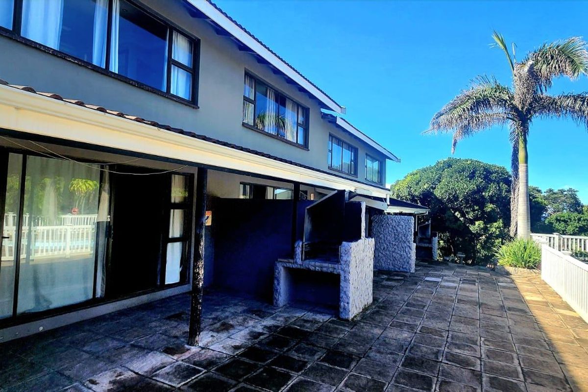 Rear exterior view featuring a spacious tiled patio with a built-in braai, sliding door access, and a white balustrade overlooking the garden area.