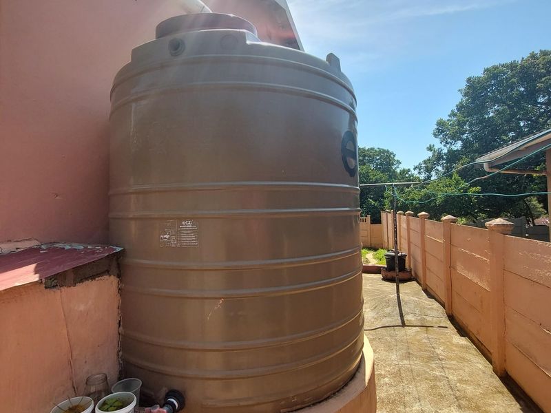 Rear exterior area featuring a large water storage tank mounted on a concrete base, adjacent to a boundary wall and paved walkway.