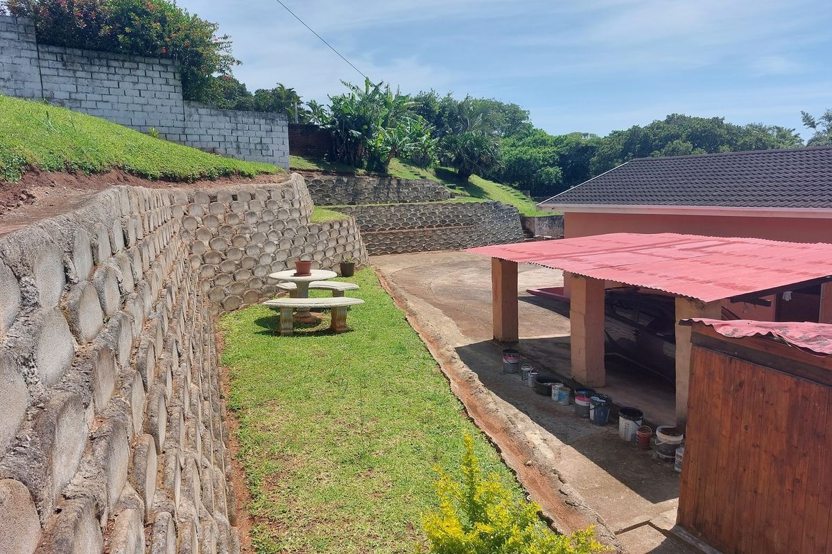 Rear exterior view featuring tiered concrete retaining walls, a grassed garden area, and a covered carport structure with a red roof.