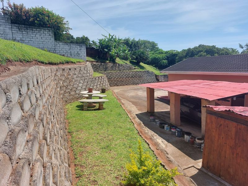 Rear exterior view featuring tiered concrete retaining walls, a grassed garden area, and a covered carport structure with a red roof.