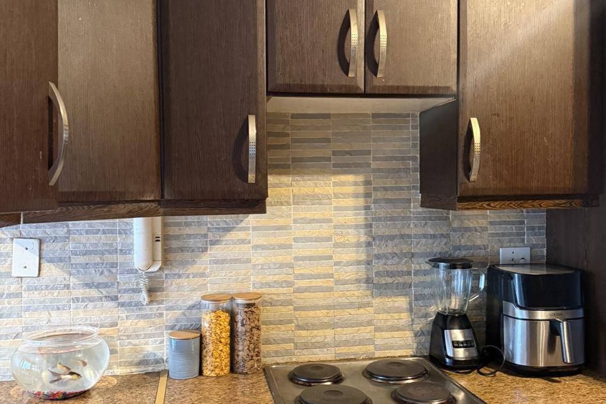 A compact kitchen area featuring dark wood-finish cabinetry, a stone-look laminate countertop, and a built-in electric hob and oven with a tiled backsplash.