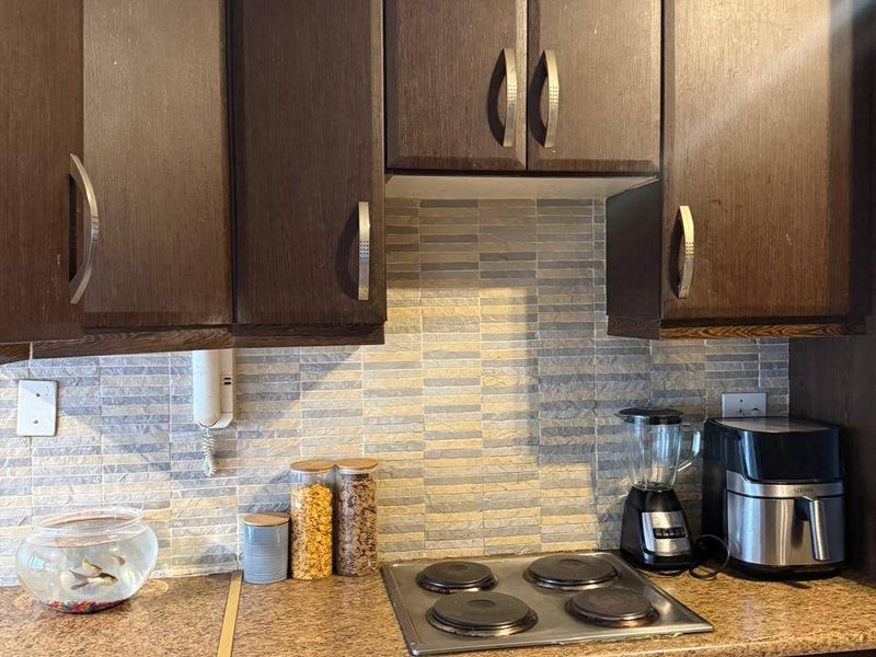 A compact kitchen area featuring dark wood-finish cabinetry, a stone-look laminate countertop, and a built-in electric hob and oven with a tiled backsplash.