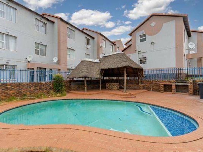Communal swimming pool area featuring a thatched lapa, built-in braai facility, and brick paving, set within a townhouse complex.