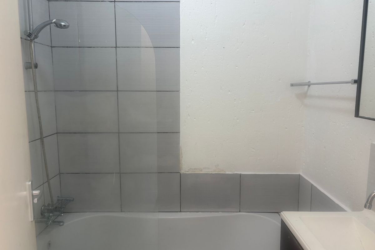 A functional bathroom featuring a bathtub with a glass shower screen, tiled splashback, and a vanity unit, requiring some cosmetic wall repairs.