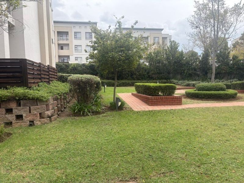 A well-maintained communal garden area featuring manicured hedges, brick pathways, and a retaining wall, set within a residential apartment complex.