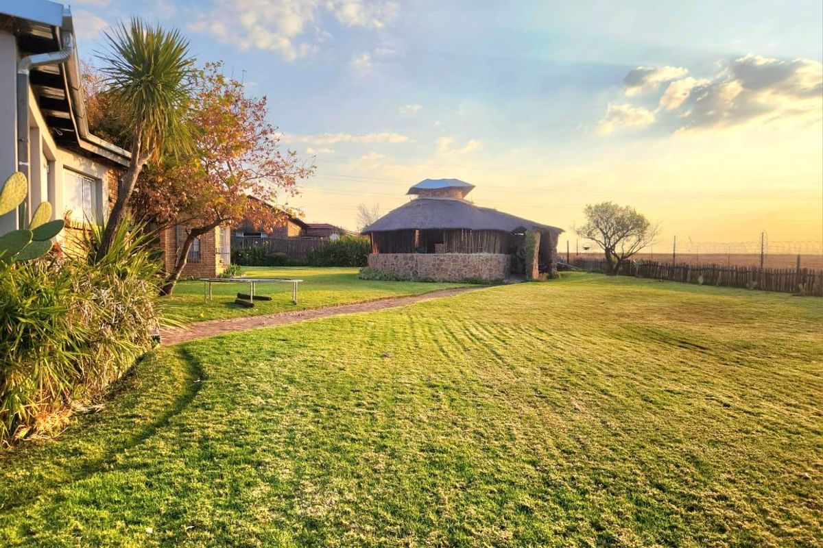 A spacious rear garden featuring a traditional thatched lapa with stone masonry, a brick pathway, and well-maintained lawn area at sunset.