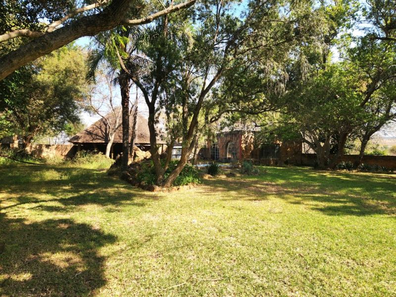 A spacious, tree-lined garden featuring a mature lawn, stone boundary walls, and a rustic thatched-roof structure in the background.