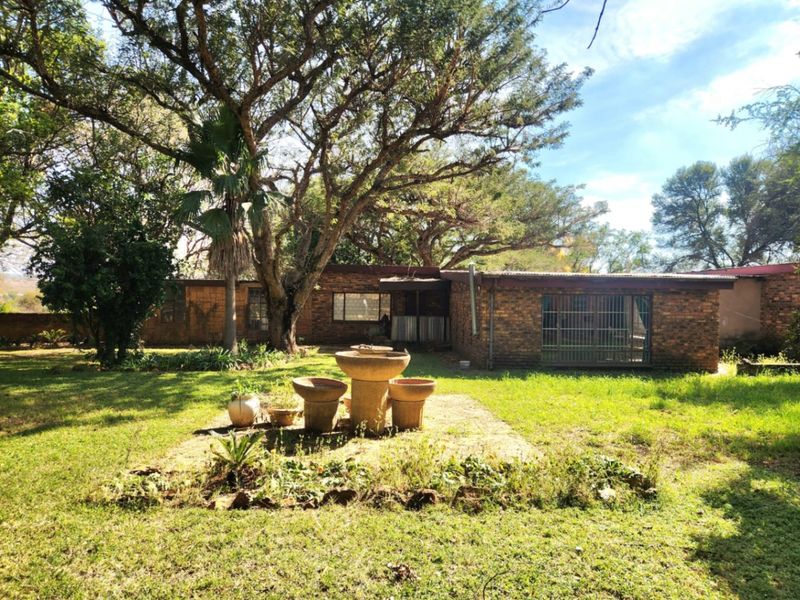 Rear view of a brick farmhouse featuring a large garden, mature trees, and a central stone fountain feature.