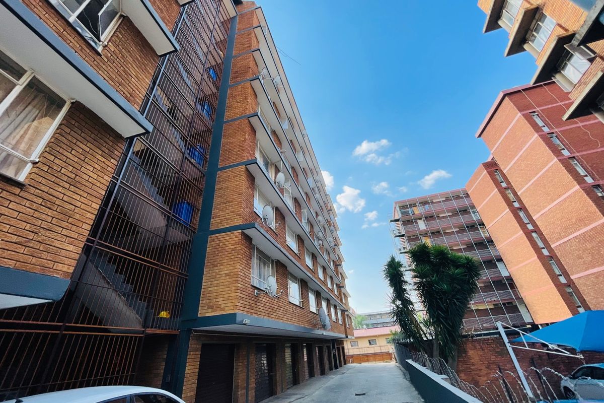 Exterior view of a multi-story brick apartment building featuring external stairwells, security fencing, and ground-level parking access.