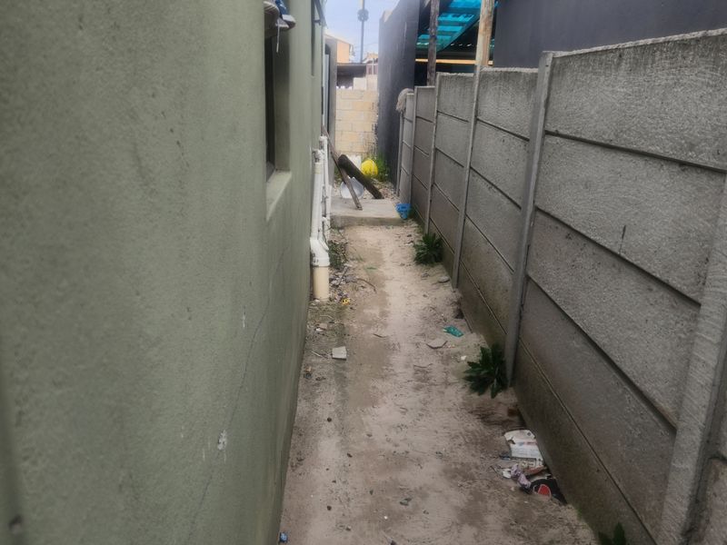 A narrow side passage between the house exterior and a precast concrete boundary wall, featuring unpaved ground and exposed external plumbing.