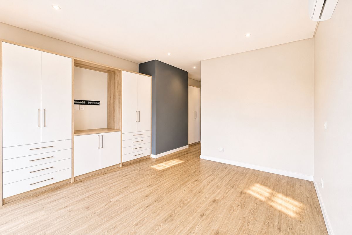 A bright, modern bedroom featuring light wood-look laminate flooring, extensive built-in cabinetry with integrated shelving, and a wall-mounted air conditioning unit.