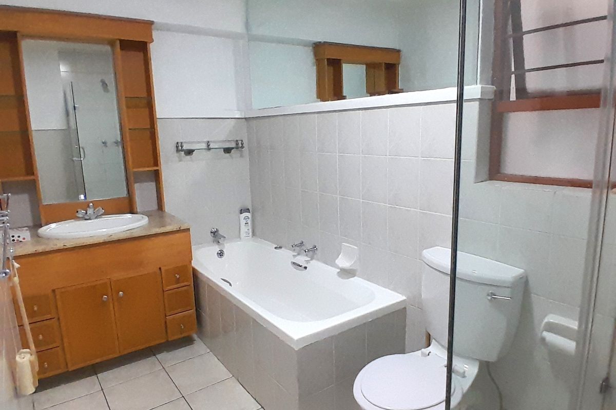 A functional bathroom featuring a built-in wooden vanity, bathtub, toilet, and a glass-enclosed shower area with neutral wall and floor tiling.