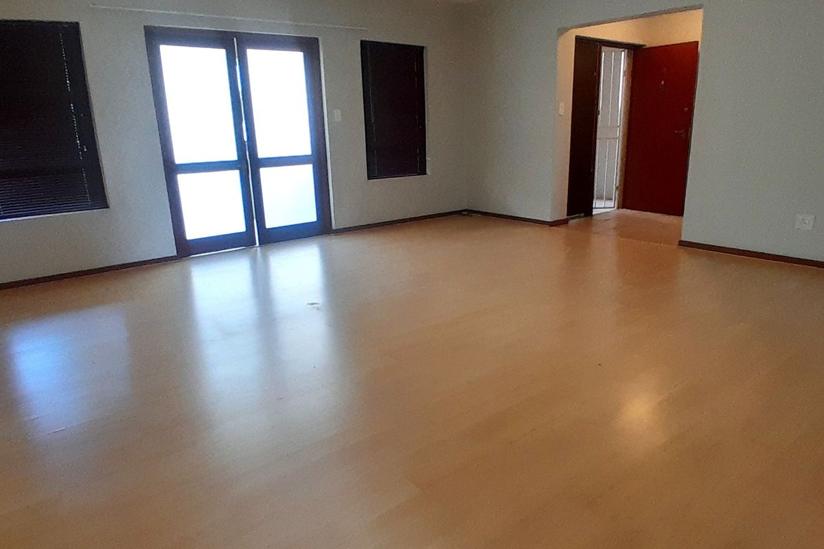 A spacious, empty living room featuring light-toned laminate flooring, double glass doors with blinds, and neutral wall finishes.