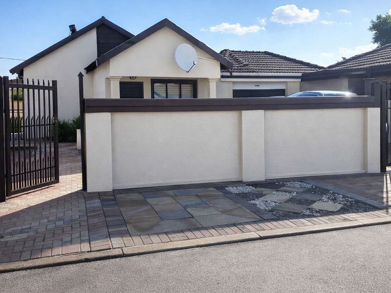 Front view of the property featuring a paved driveway, secure metal gate, and a well-maintained boundary wall with a tiled roof facade.