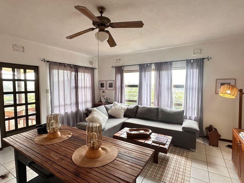 A bright living room featuring tiled flooring, a ceiling fan, and large windows that provide natural light and access to an outdoor area.