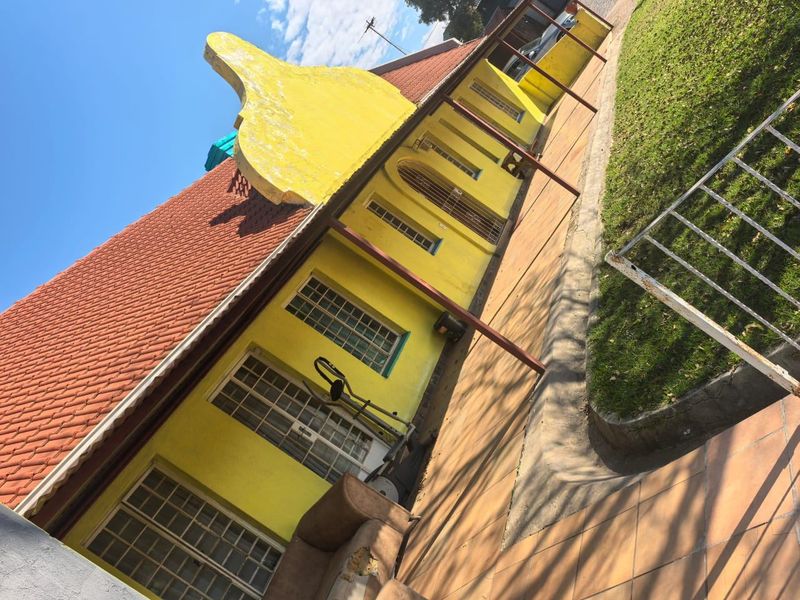 Rear exterior view of a commercial building featuring a gabled roof, yellow facade, security-barred windows, and a paved patio area adjacent to a lawn.