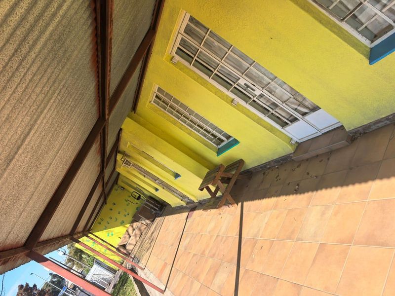A covered outdoor patio area featuring tiled flooring and yellow exterior walls with multiple windows, providing a functional transition space for the commercial property.