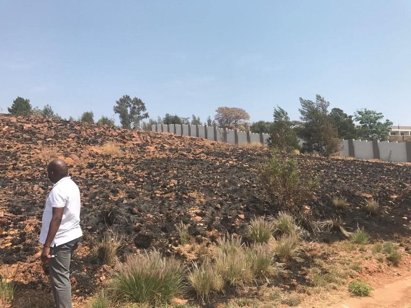A view of a sloped vacant residential plot featuring a perimeter wall and signs of recent fire damage to the ground cover.