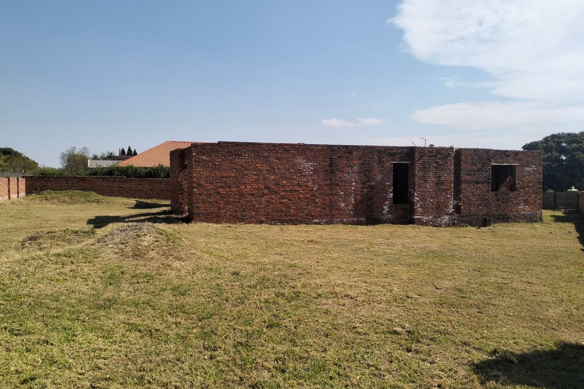 A view of an unfinished brick structure situated on a large plot of land, showing the building shell and surrounding yard.