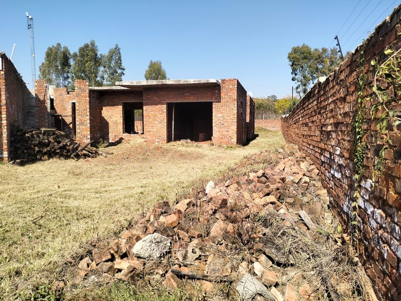 An incomplete residential structure showing exposed brickwork and concrete slab roofing, situated on a large plot with a brick boundary wall.
