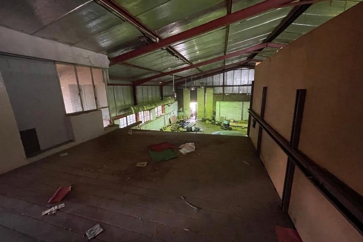 An industrial mezzanine level overlooking a large warehouse space, featuring exposed steel roof trusses and significant wear requiring refurbishment.
