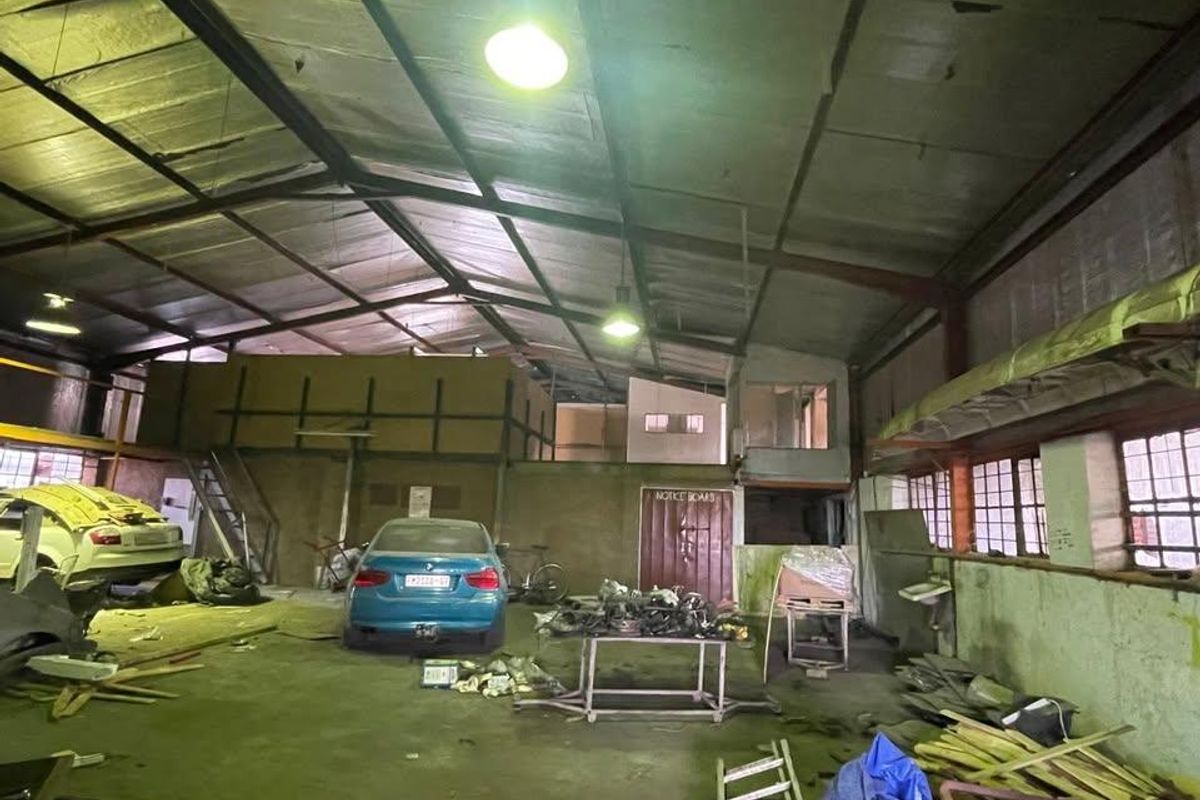 Spacious industrial garage or workshop area featuring a mezzanine level, high steel-truss ceilings, and large windows, requiring significant maintenance and clearing.