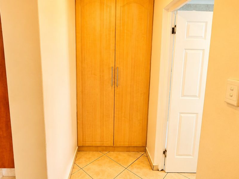 A functional hallway featuring built-in wooden cupboards, tiled flooring, and access to adjacent rooms, including a glimpse into the kitchen area.