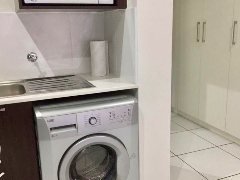A compact laundry area featuring built-in cabinetry, a stainless steel sink, and tiled flooring, adjacent to a hallway with built-in storage cupboards.