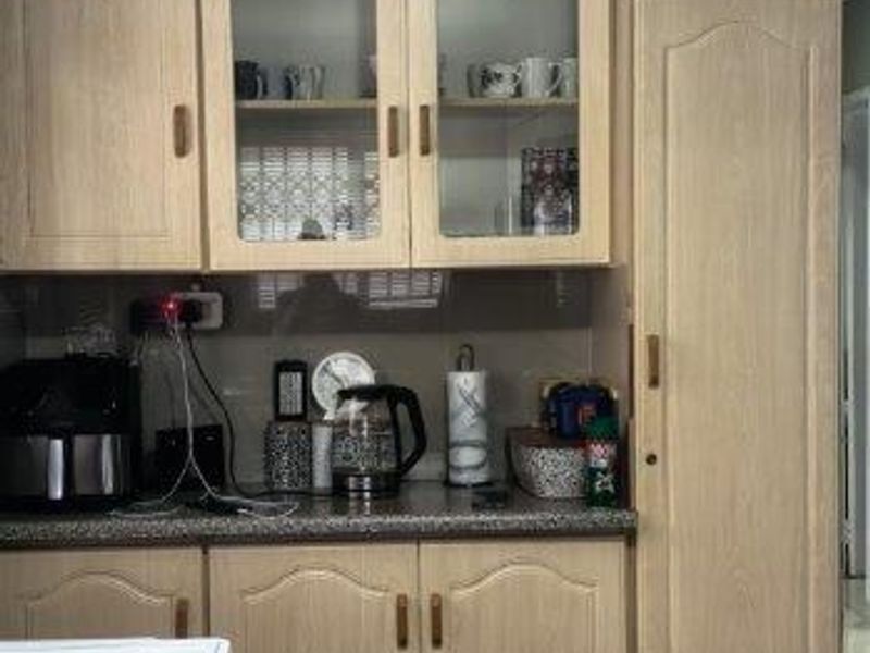 A functional kitchen area featuring light wood-grain laminate cabinetry, glass-fronted display cupboards, a tall pantry unit, and tiled flooring.