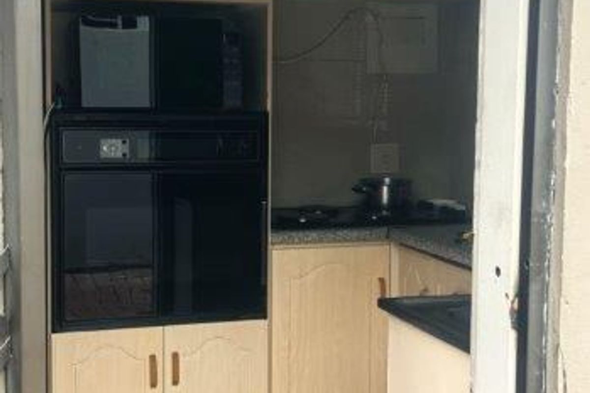 A compact kitchen featuring light wood-finish cabinetry, built-in oven and microwave, dark countertops, and tiled flooring, viewed through a doorway.