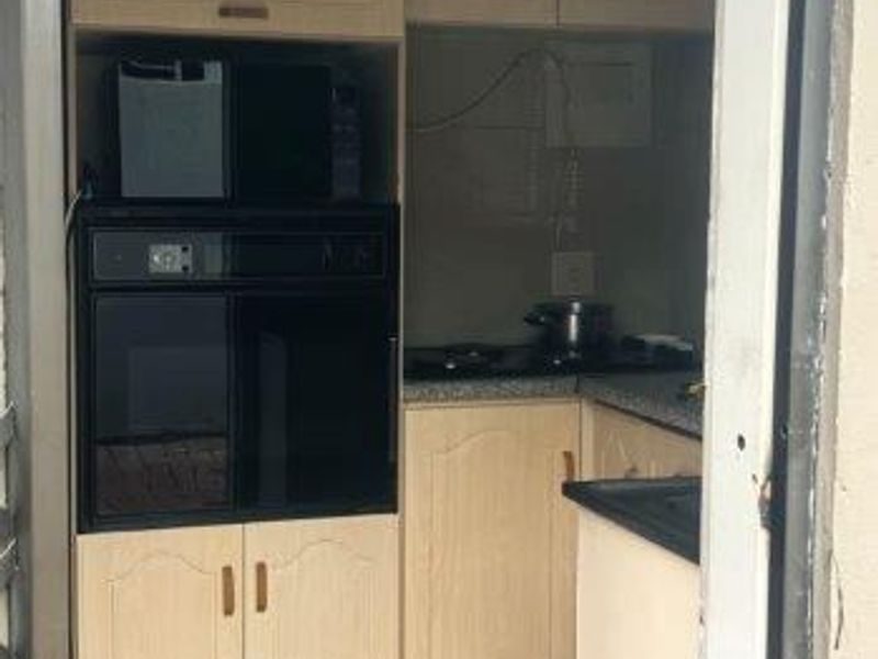 A compact kitchen featuring light wood-finish cabinetry, built-in oven and microwave, dark countertops, and tiled flooring, viewed through a doorway.