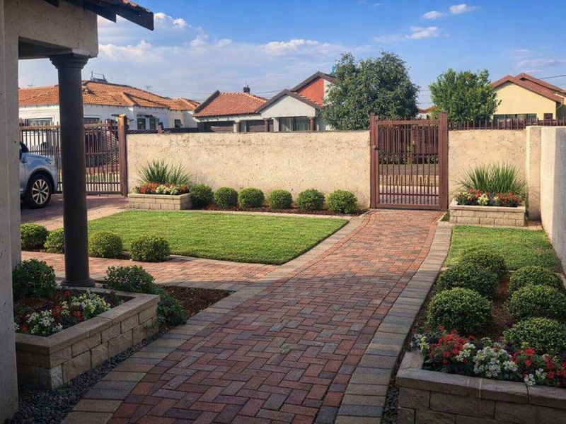 A well-maintained front entrance area featuring a brick paved walkway, manicured lawn, raised garden beds, and secure perimeter walling with a metal gate.