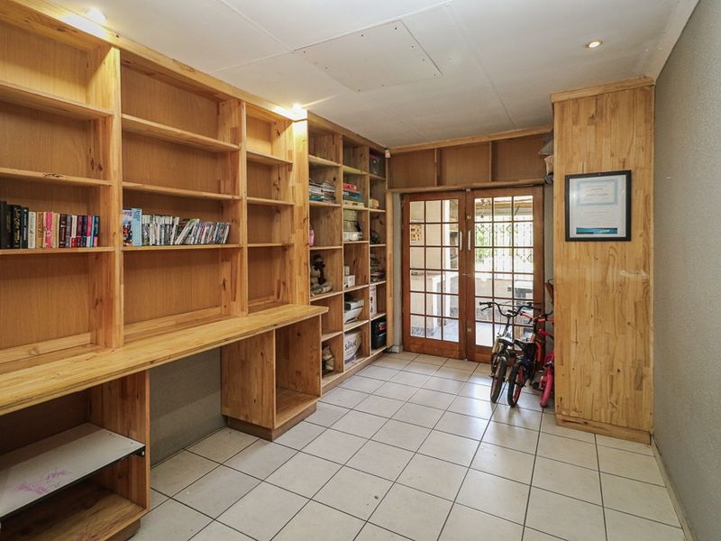 A functional home office or study area featuring extensive built-in wooden shelving, a desk workspace, tiled flooring, and direct access to an outdoor patio.