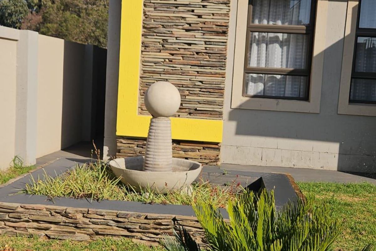 A well-maintained rear garden area featuring a stone-clad facade, a decorative concrete water feature, and established landscaping.