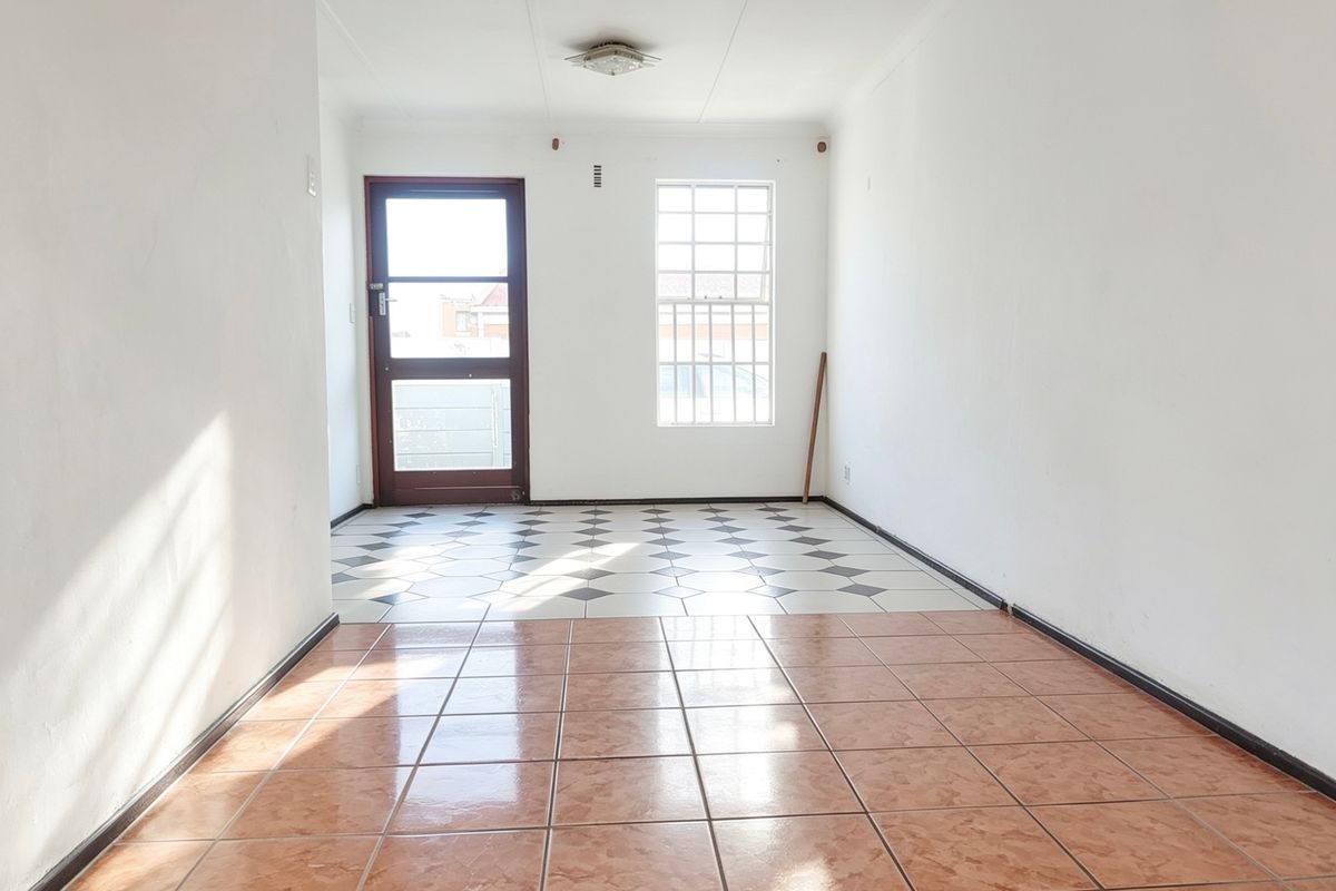 An empty living area featuring dual-tone tiled flooring, a security door, and a window with burglar bars, offering a blank canvas for renovation.